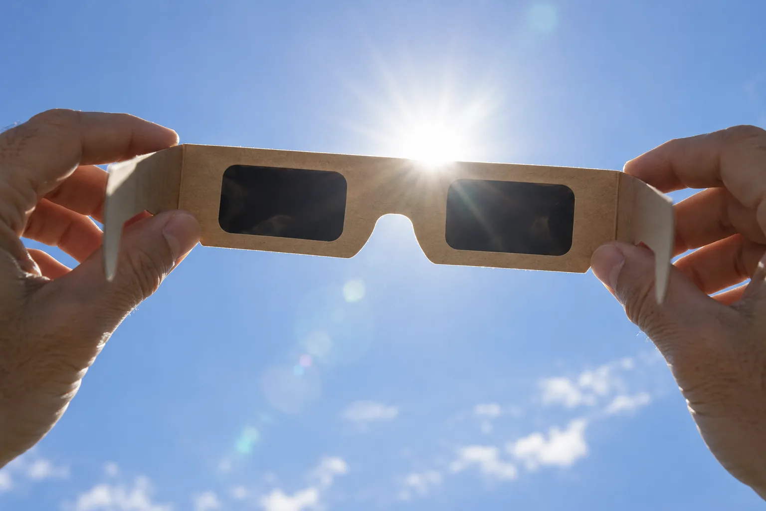 Hands holding cardboard solar eclipse glasses up toward the bright sun